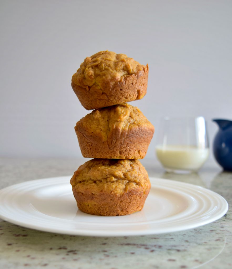 3 muffins stacked on top of each other on a white plate, glass of milk and blue glass in background