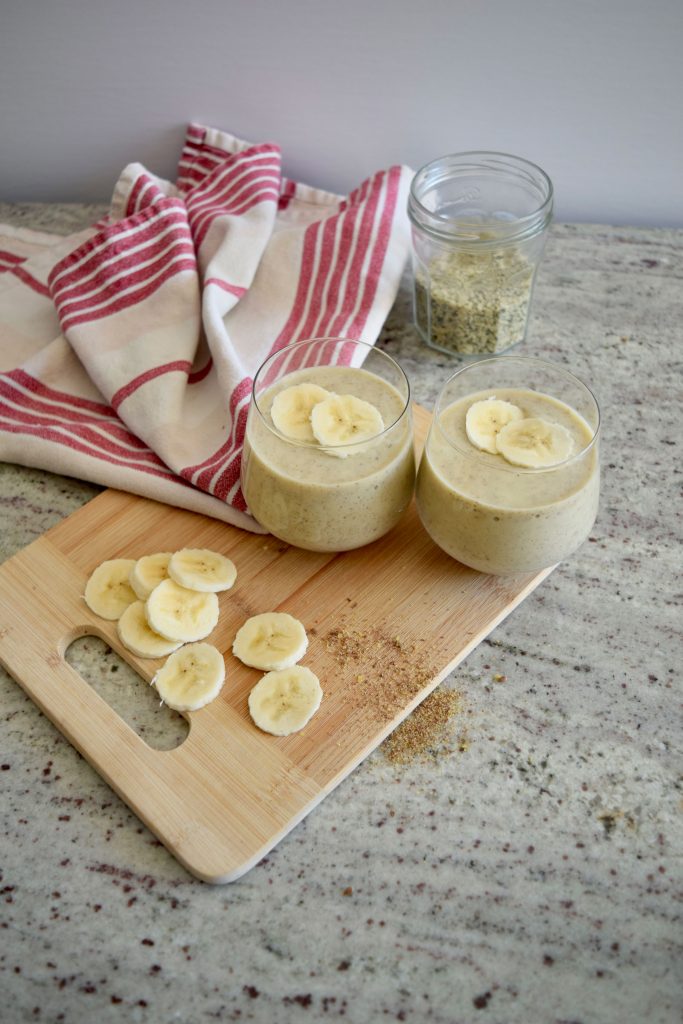 two smoothies on a wooden cutting board, banana slices to the side and a napkin and jar in the distance