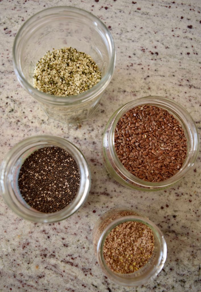 4 jars containing hemp hearts, chia seeds, flax seeds and ground flax on a marble countertop