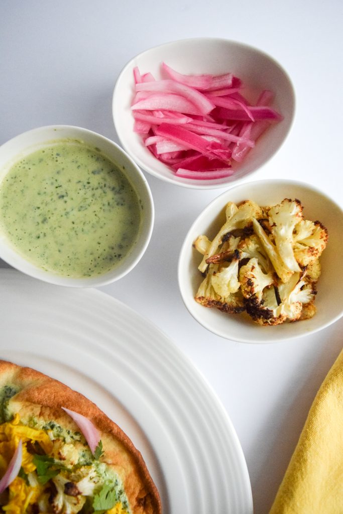 3 small white bowls containing: cilantro yogurt sauce, roasted cauliflower and pickled red onion