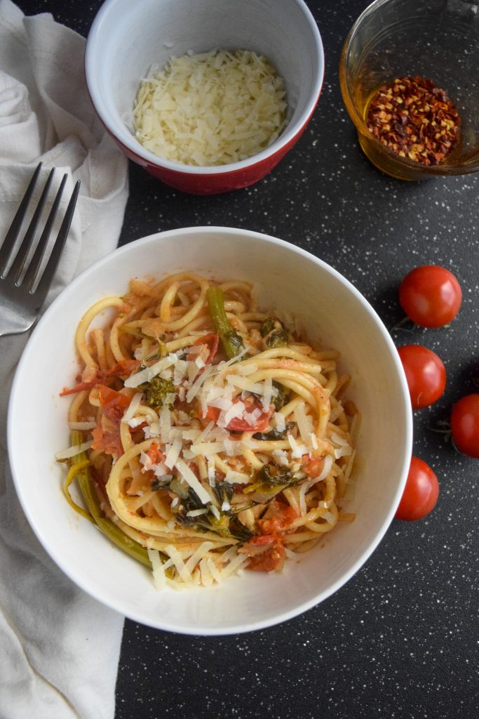 tomato and rapini pasta topped with cheese in a white bowl, surrounded by smaller bowls, tomatoes and a fork