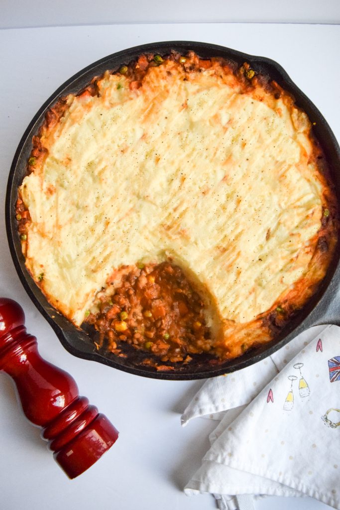mushroom & lentil shepherd's pie in a cast-iron skillet on a white backdrop, red pepper shaker to the left, white tea towel on the right