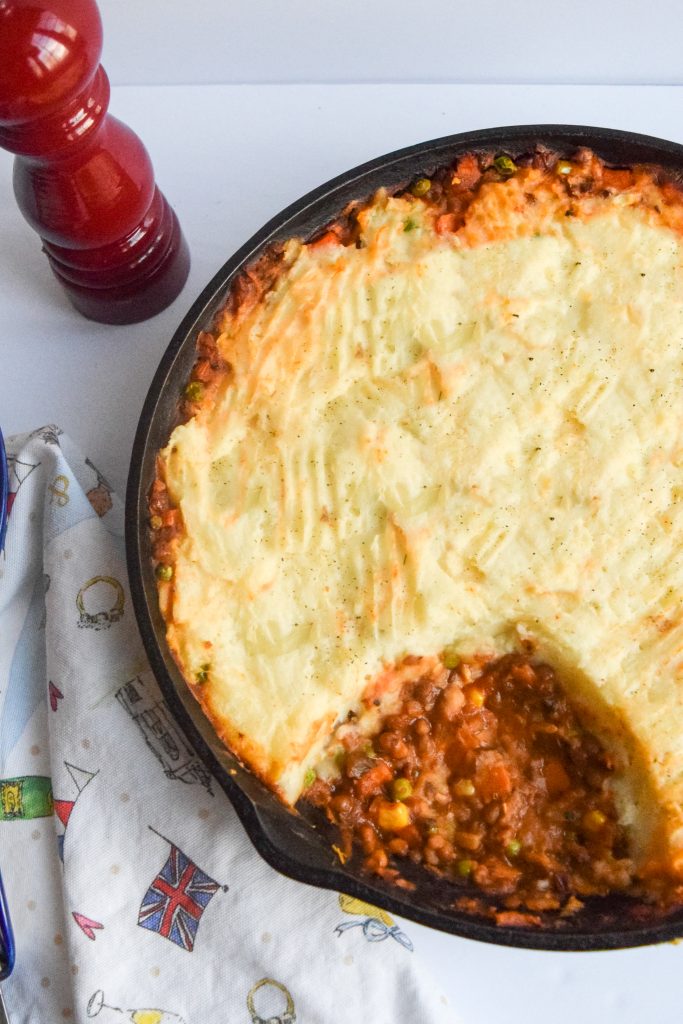 mushroom & lentil shepherd's pie in a cast-iron skillet on a white backdrop, red pepper shaker to the left, white tea towel on the right