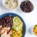 Blue bowl on a white backdrop filled with salmon, black beans, guacamole and mango salsa. Surrounded by a fork, 2 small bowls and an avocado.