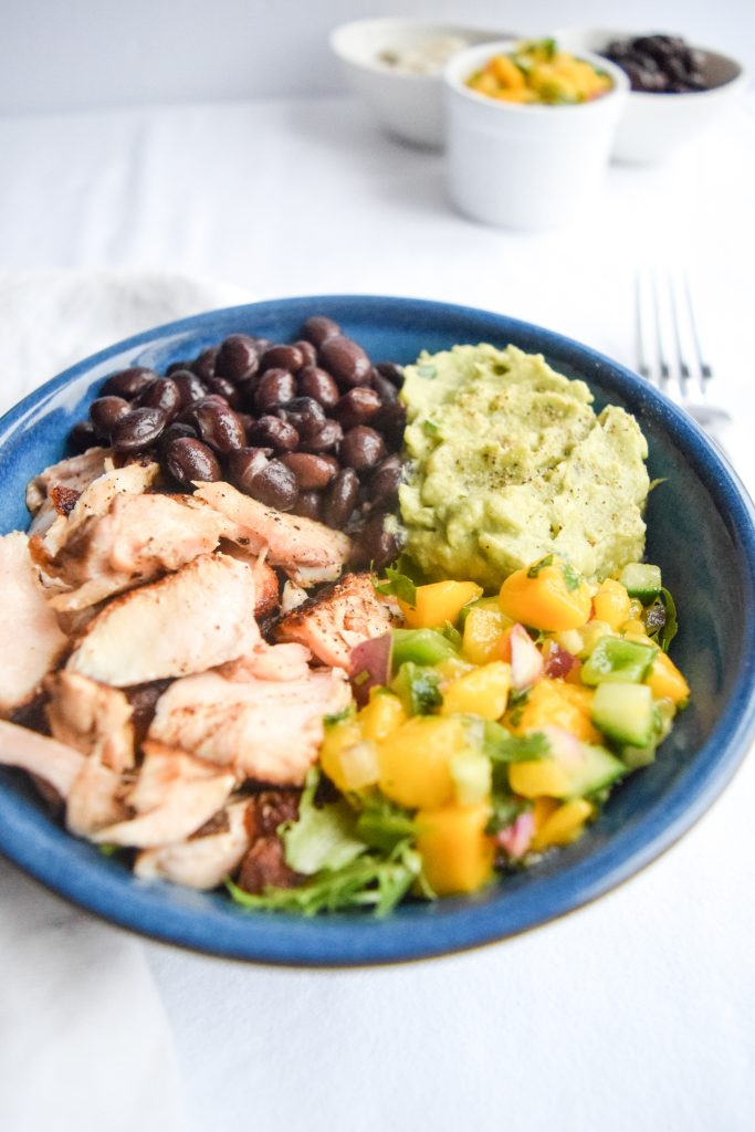 Blue bowl on a white backdrop filled with salmon, black beans, guacamole and mango salsa. 3 small bowls in the distance.