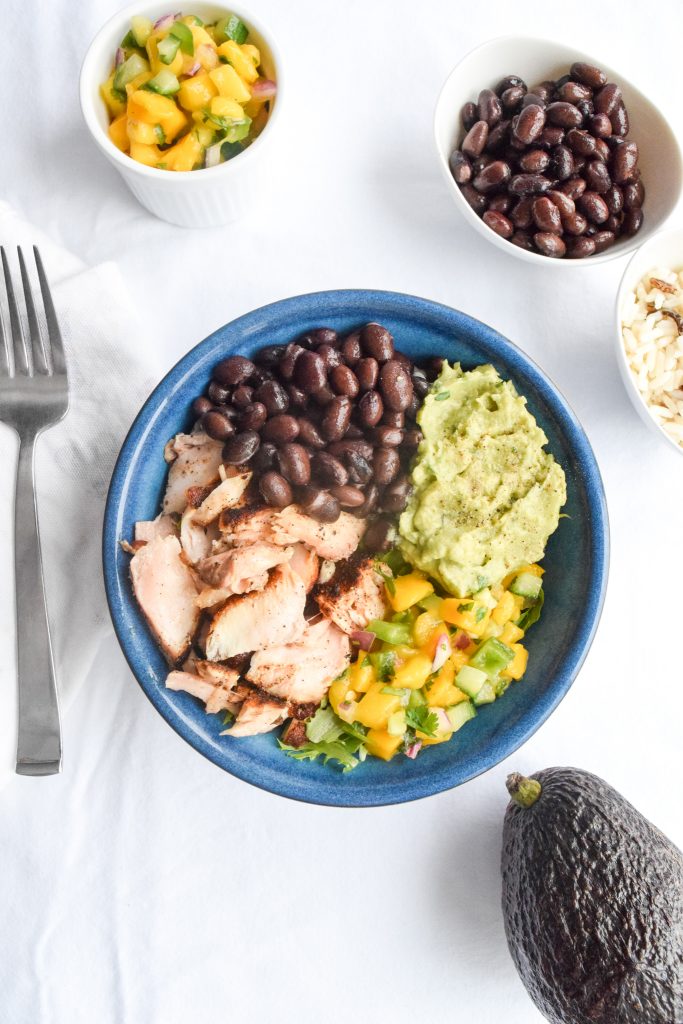 Blue bowl on a white backdrop filled with salmon, black beans, guacamole and mango salsa. Surrounded by a fork, 2 small bowls and an avocado.
