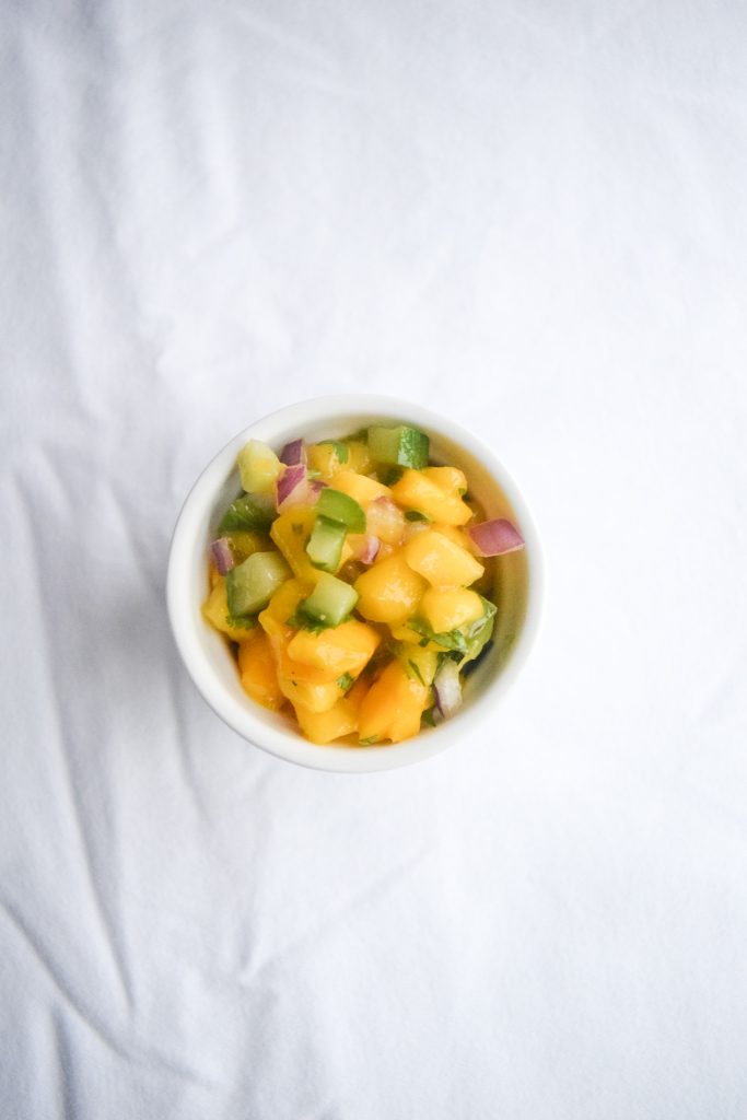 Small white bowl filled with mango salsa on a white backdrop.