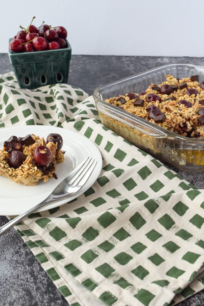 white plate with a piece of baked oatmeal on it, to the right a glass dish with the remainder of the baked oats and cherries in the distance