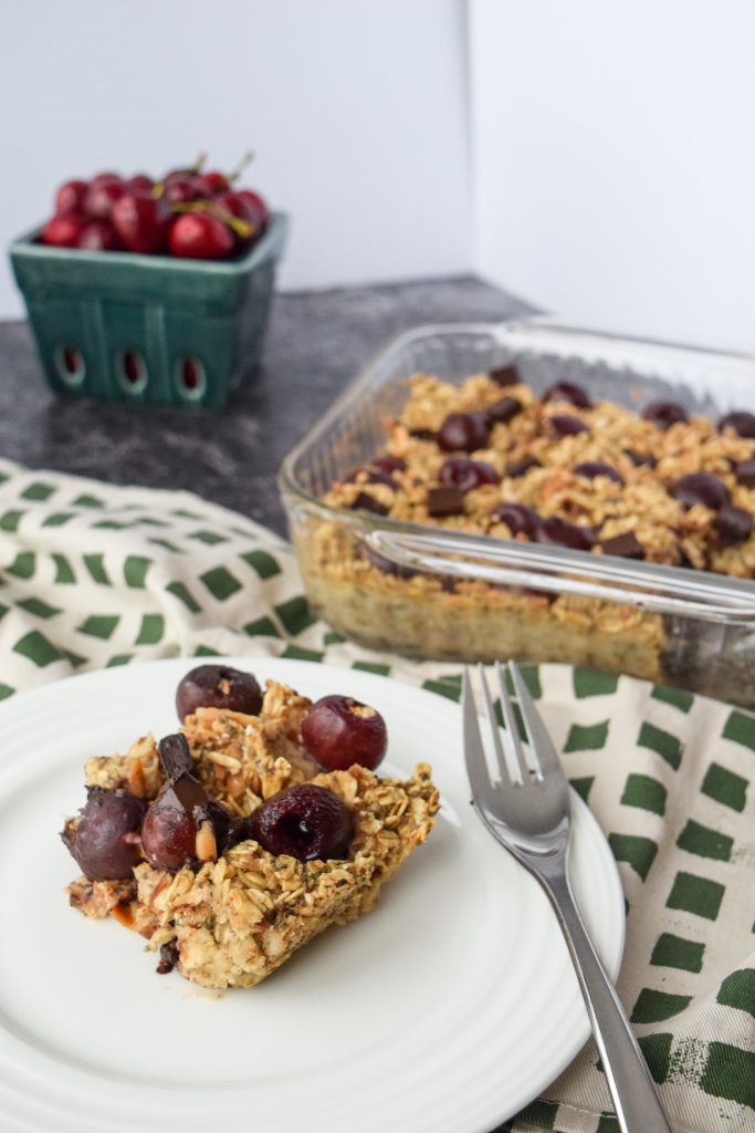 white plate with a piece of baked oatmeal on it, to the right a glass dish with the remainder of the baked oats and cherries in the distance