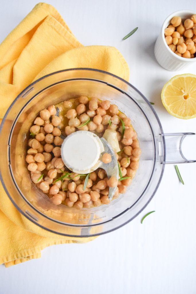 Food processor filled with chickpeas, garlic and herbs. Surrounded by a yellow napkin, half a lemon and a white bowl with chickpeas