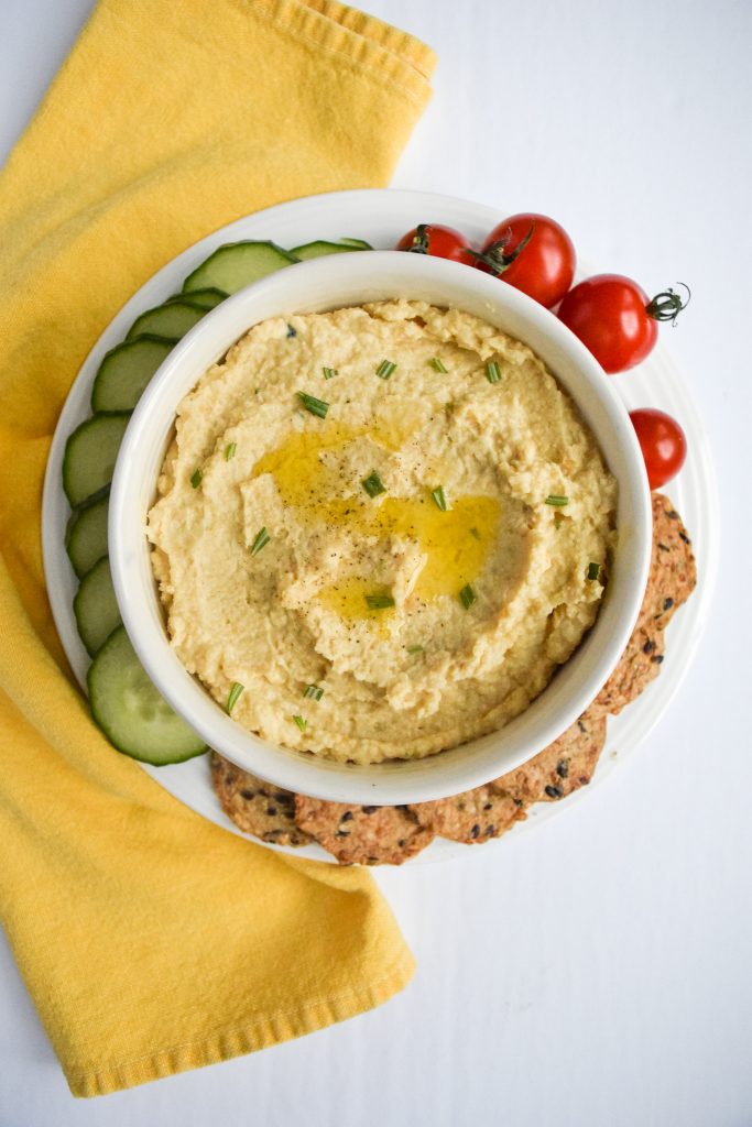 white bowl of hummus topped with fresh herbs and oil, surrounded by cucumbers, cherry tomatoes and crackers, and a yellow napkin