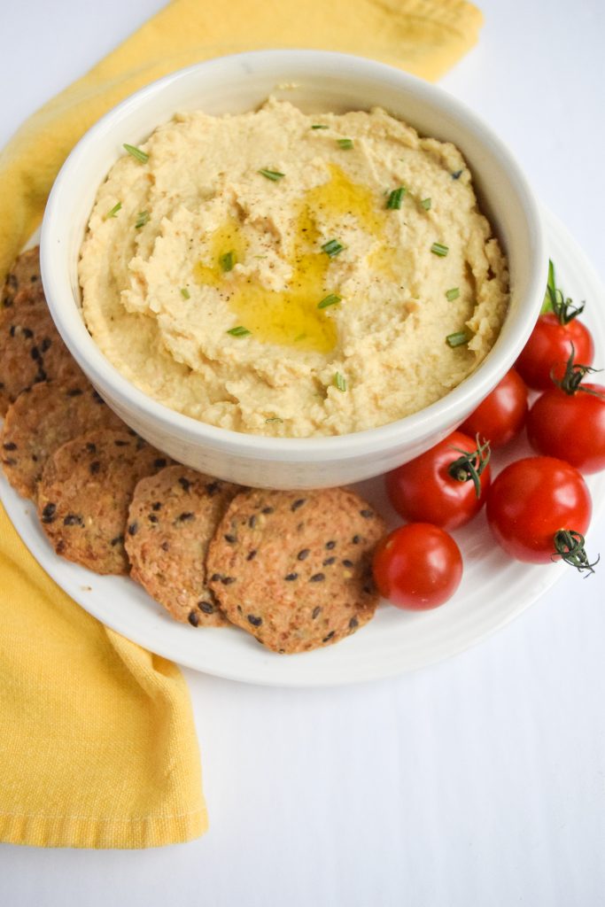 white bowl of hummus surrounded by crackers and cherry tomatoes, yellow napkin to left