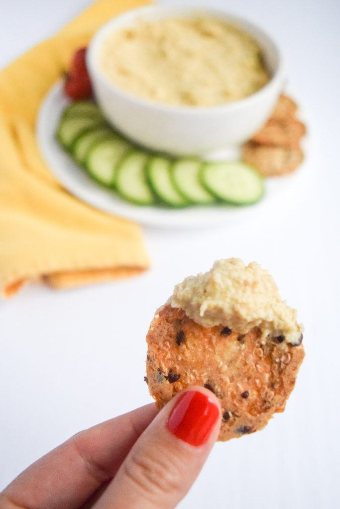 two fingers holding up a cracker with hummus on it. Bowl of hummus surrounded by veggies and a yellow napkin in the back