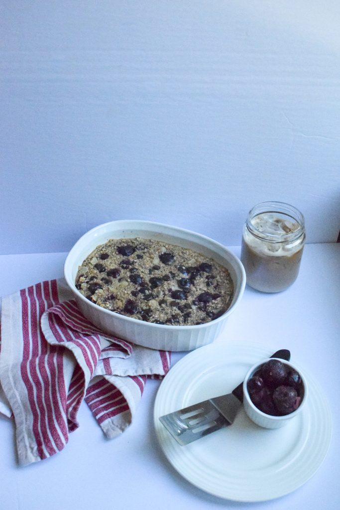 white casserole dish filled with baked oatmeal, topped with cherries and dark chocolate, surrounded by a small bowl of cherry and a jar of iced coffee