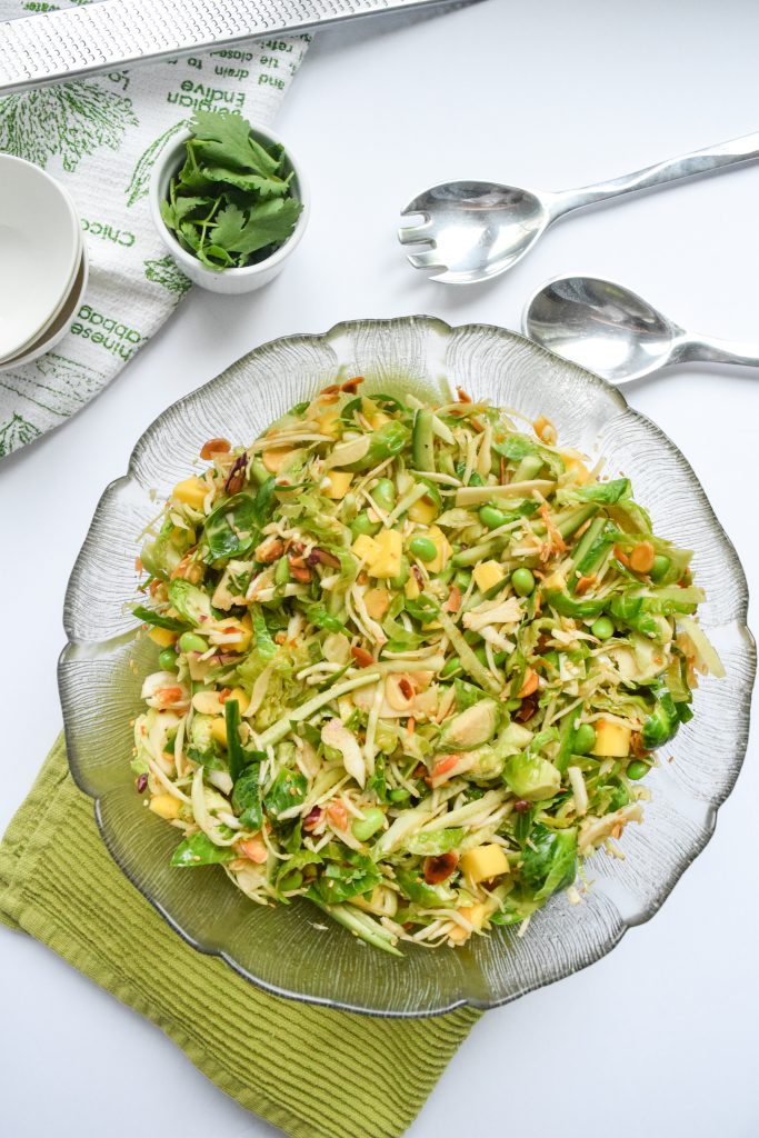 Green slaw in a glass bowl, atop a green napkin. Surrounded by white bowls, silver salad tongs