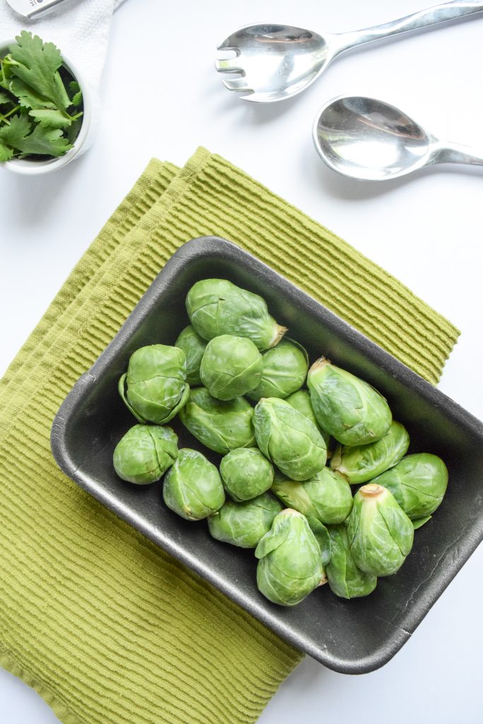 black tray filled with brussels sprouts on a green tea towel, surrounded by a white bowl filled with cilantro and silver salad tongs
