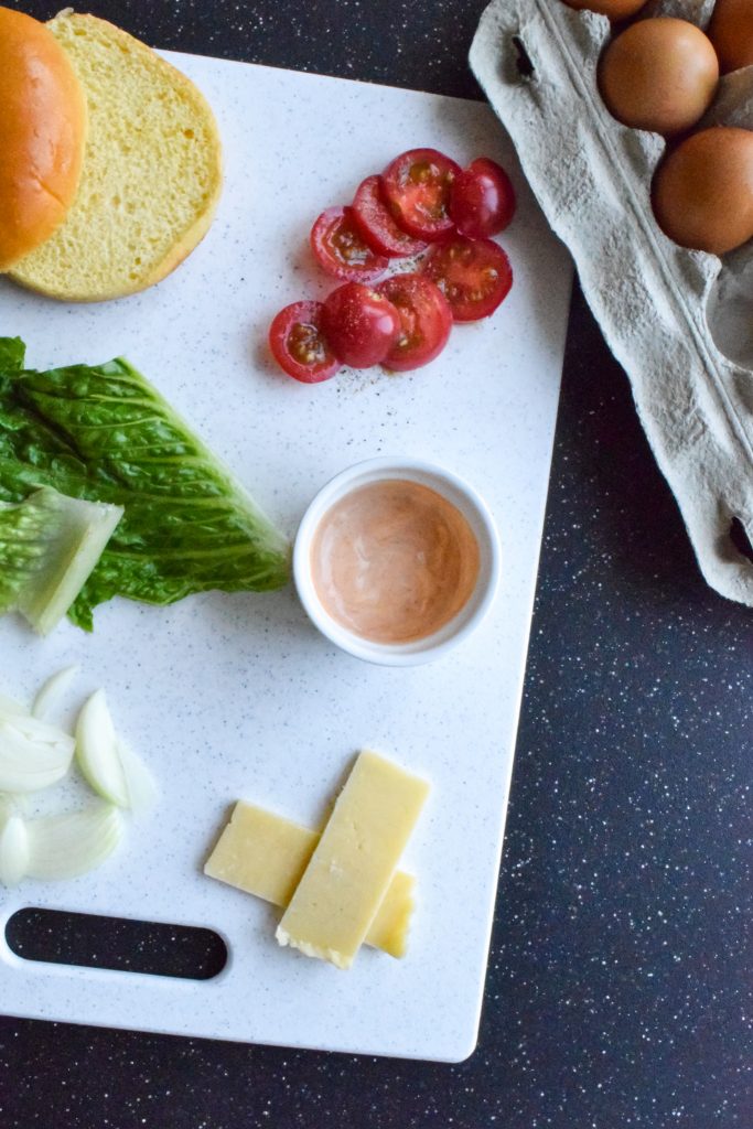 Black backdrop with a white cutting board, filled with cheese slices, spicy mayo, onion, lettuce, brioche bun and sliced tomatoes. Egg carton in upper right corner.