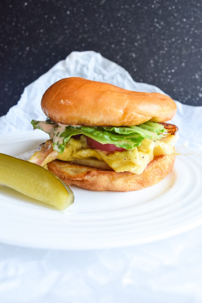 White plate with a breakfast sandwich on brioche bun, sliced pickle on side. Atop parchment paper with a white background.