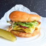 White plate with a breakfast sandwich on brioche bun, sliced pickle on side. Atop parchment paper with a white background.