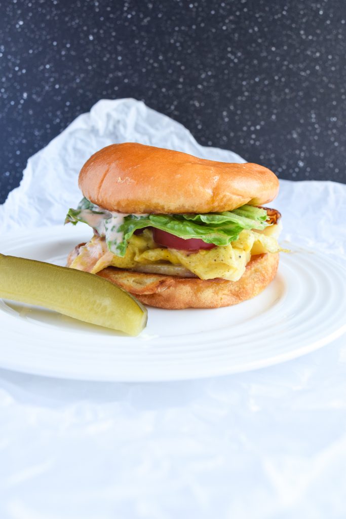 White plate with a breakfast sandwich on brioche bun, sliced pickle on side. Atop parchment paper with a white background.