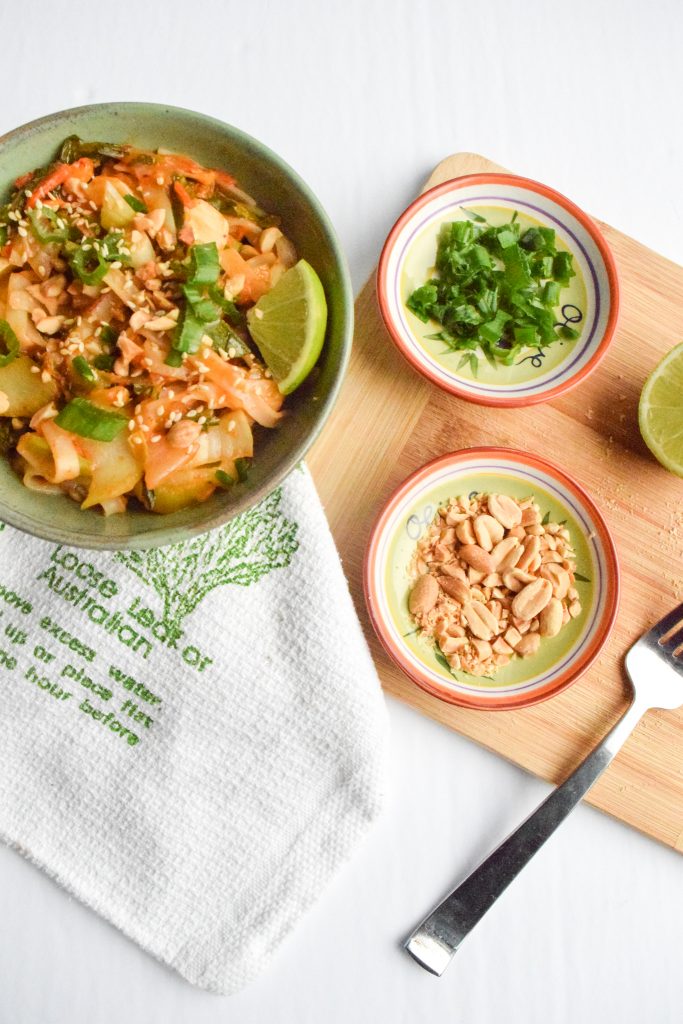 Overhead shot of a green bowl of noodles atop a white and green tea towel. Surrounded by a wooden cutting board with various small bowls, a fork and half a lime.