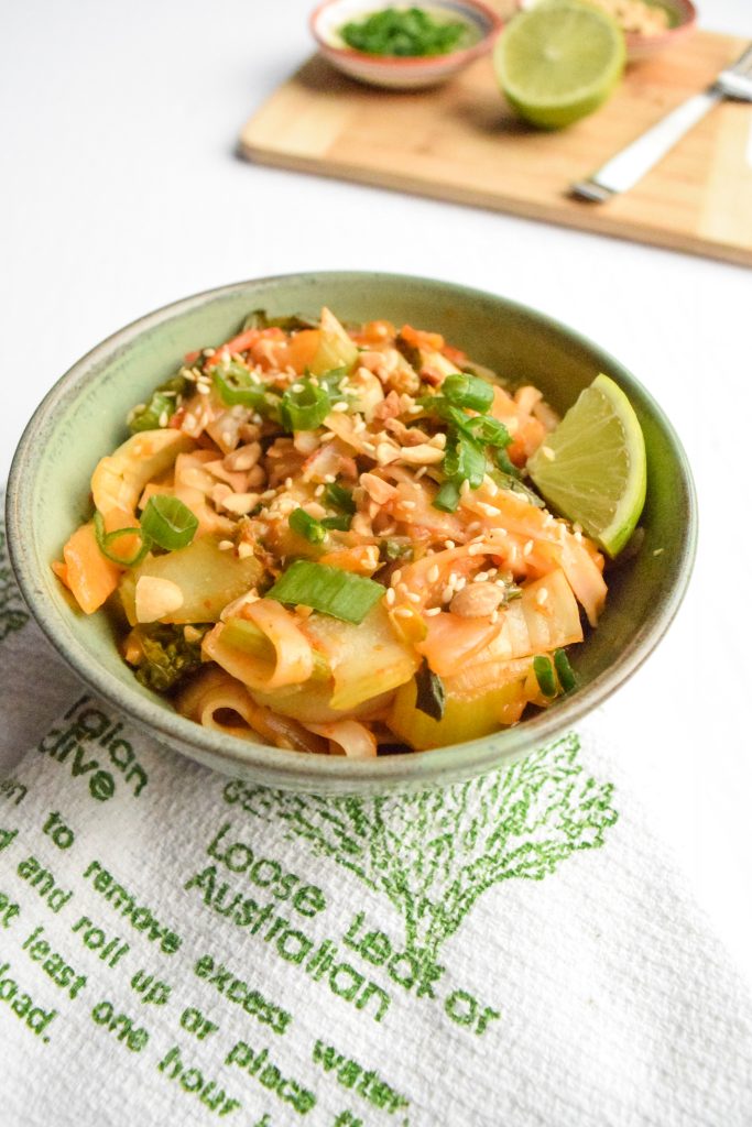 Green bowl filled with stir-fried noodles and vegetables on a white and green tea towel. In the distance, a wooden cutting board with various small bowls and ingredients.