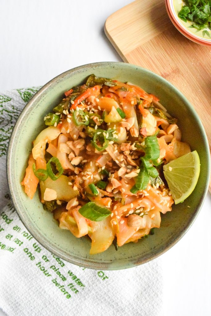 Overhead shot of a green bowl filled with stir-fried kimchi noodles and vegetables on a white and green tea towel. Wooden cutting board to the side.