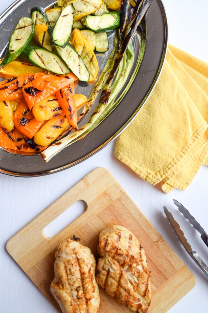 Silver tray with grilled veggies, wooden cutting board with grilled chicken, tongs and a yellow cloth napkin.