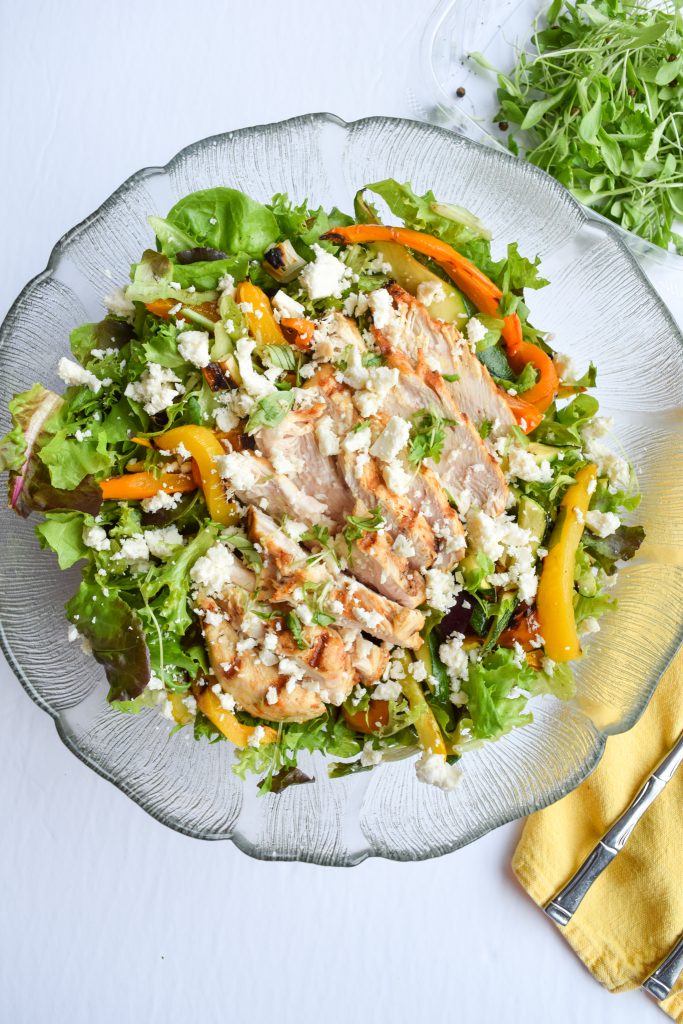 Glass salad bowl filled with salad and grilled veggies, topped with chicken and feta.