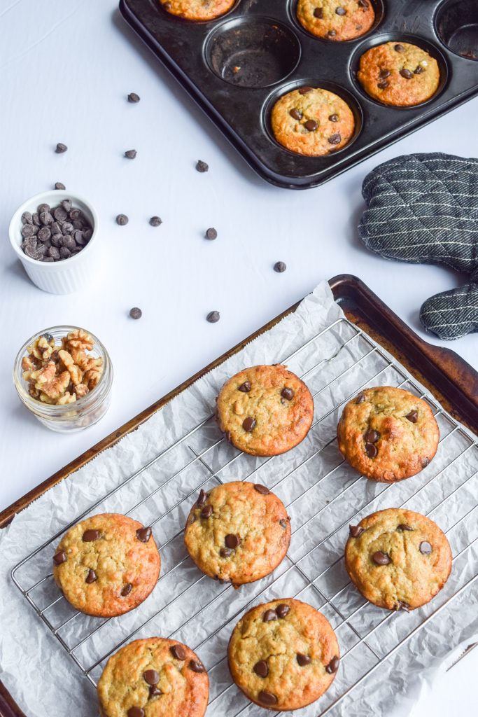 cookie sheet topped with 7 banana chocolate chip muffins, on the side, two containers, a muffin tin and a grey oven mit