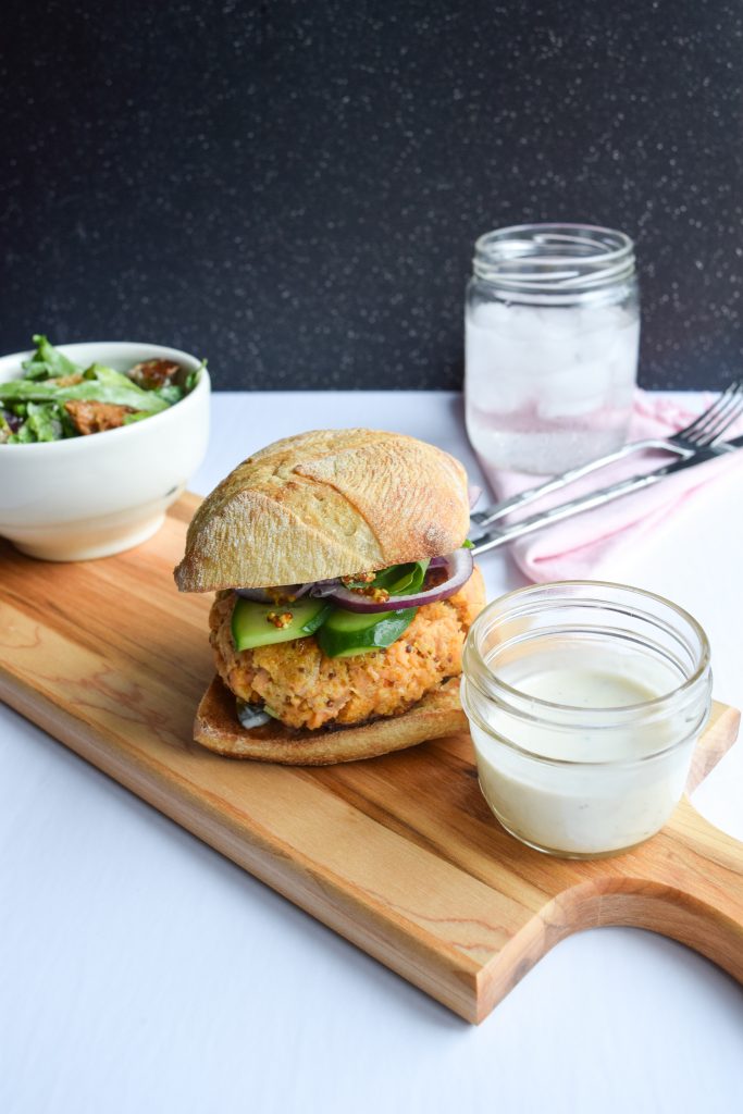 Salmon burger on a wooden plank. Surrounded by multiple bowls and jars.