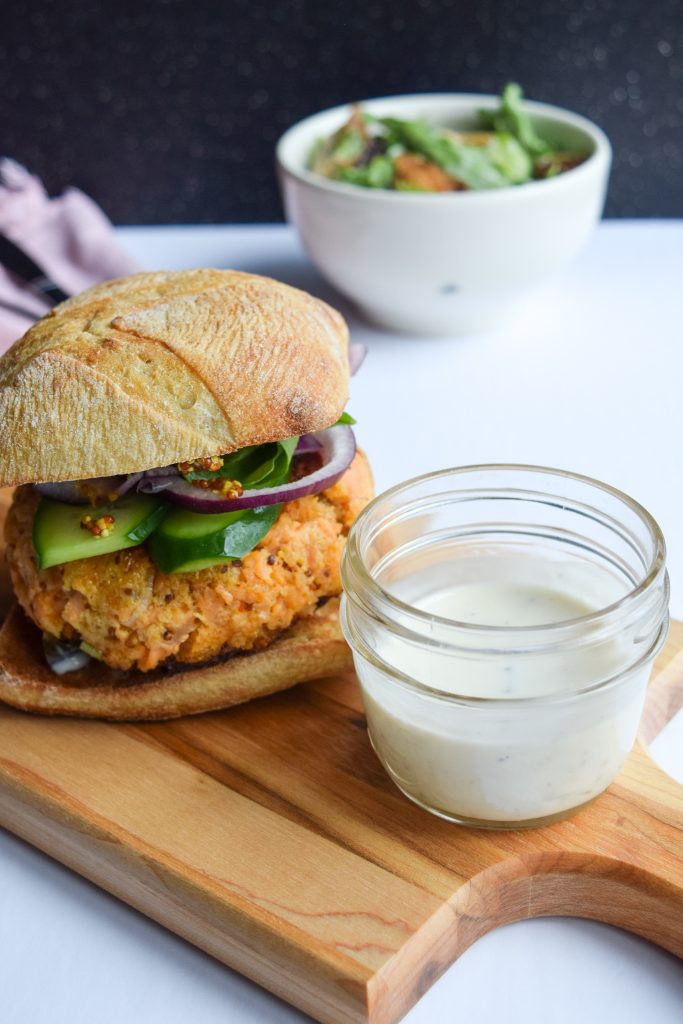 Close up shot of a salmon burger on a ciabatta bun. Next to it, a small glass jar with tartar sauce.