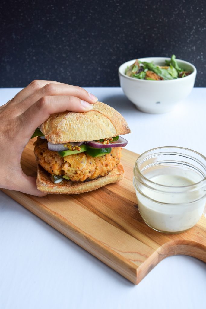 Hand grabbing a salmon burger on a ciabatta bun. Beside it, a jar of tartar sauce and a bowl of caesar salad in the distance.