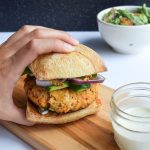 Hand grabbing a salmon burger on a ciabatta bun. Beside it, a jar of tartar sauce and a bowl of caesar salad in the distance.