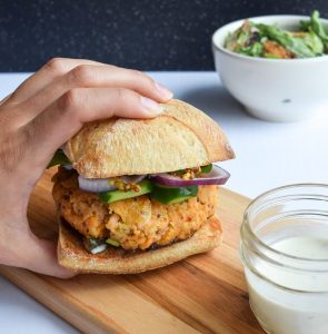 Hand grabbing a salmon burger on a ciabatta bun. Beside it, a jar of tartar sauce and a bowl of caesar salad in the distance.