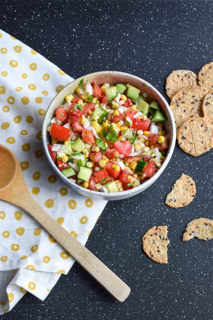 big bowl of corn salsa on a black backdrop, atop a white and yellow cloth. Beside it is a wooden spoon and crackers.