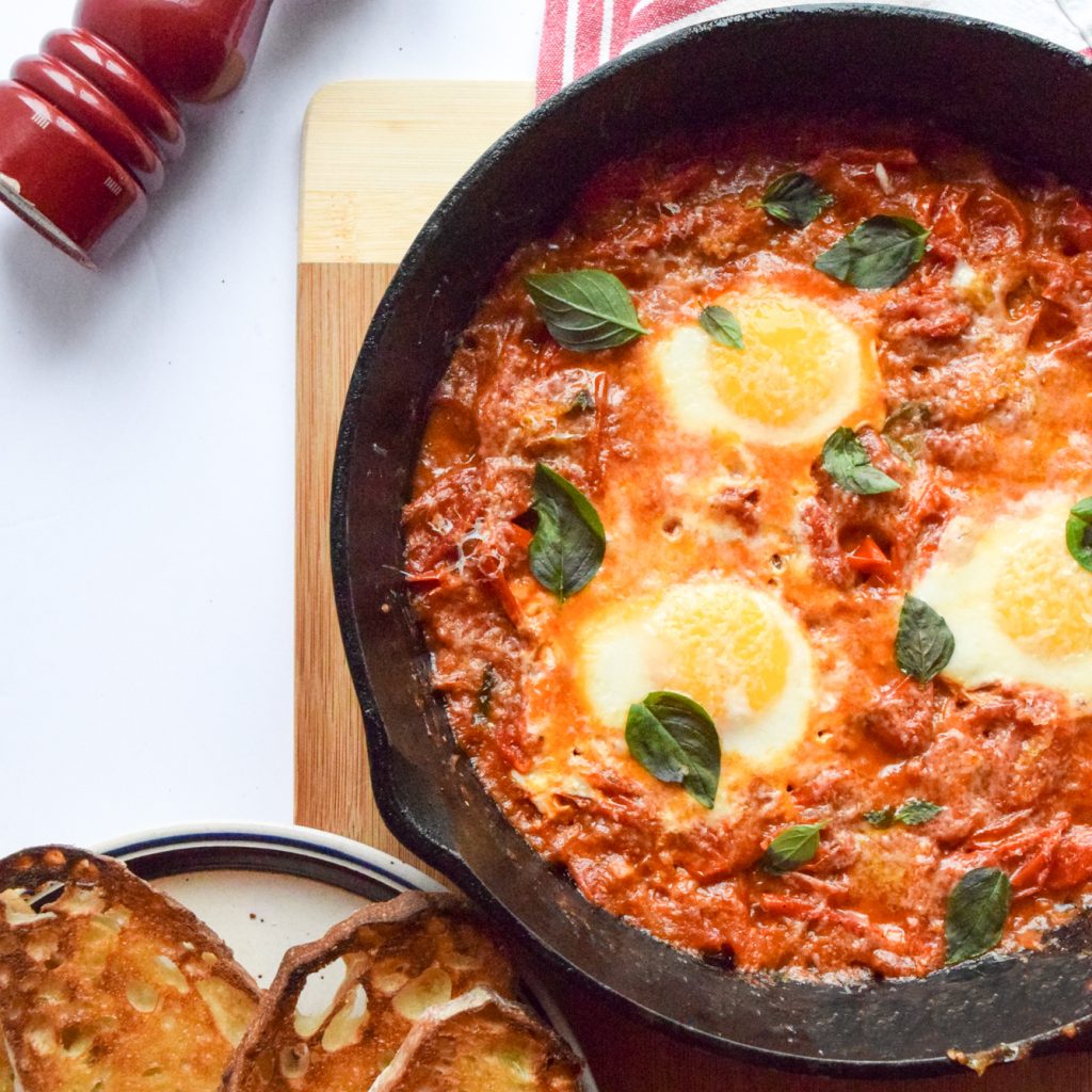 Shakshuka topped with fresh basil and parmesan cheese in a cast-iron skillet over a wooden cutting board. Aside is some toast and a red pepper grinder.