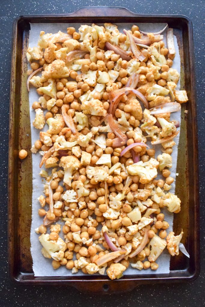Baking sheet lined with parchment paper, filled with raw, seasoned cauliflower, chickpeas and onions