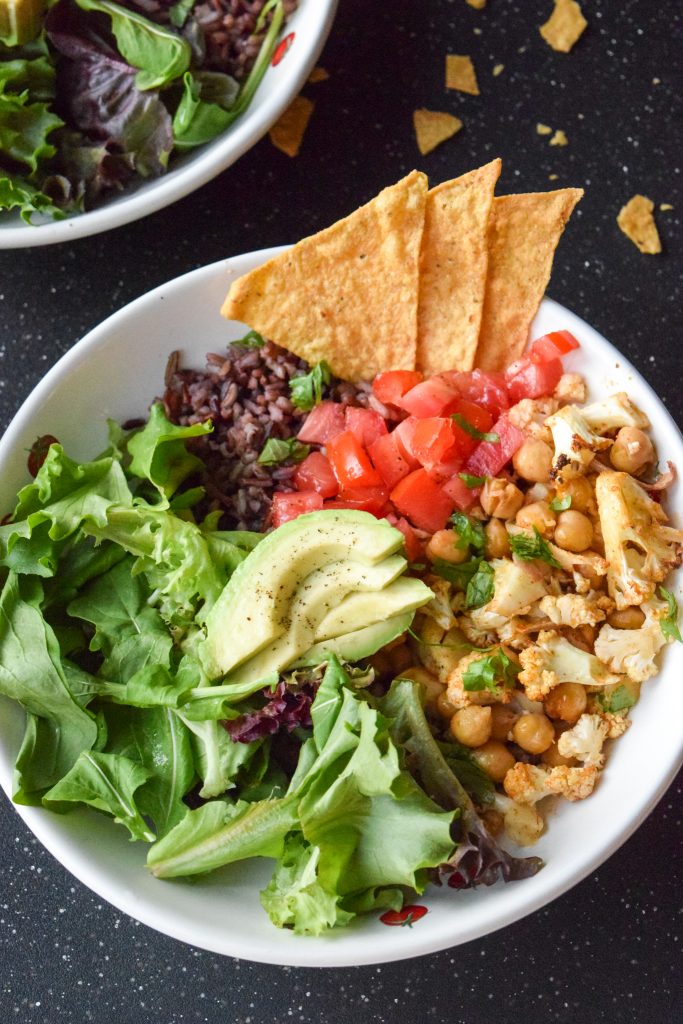 Close-up shot of a white bowl filled with lettuce, purple rice, fresh salsa, avocado and roasted taco spice cauliflower, chickpeas and onions