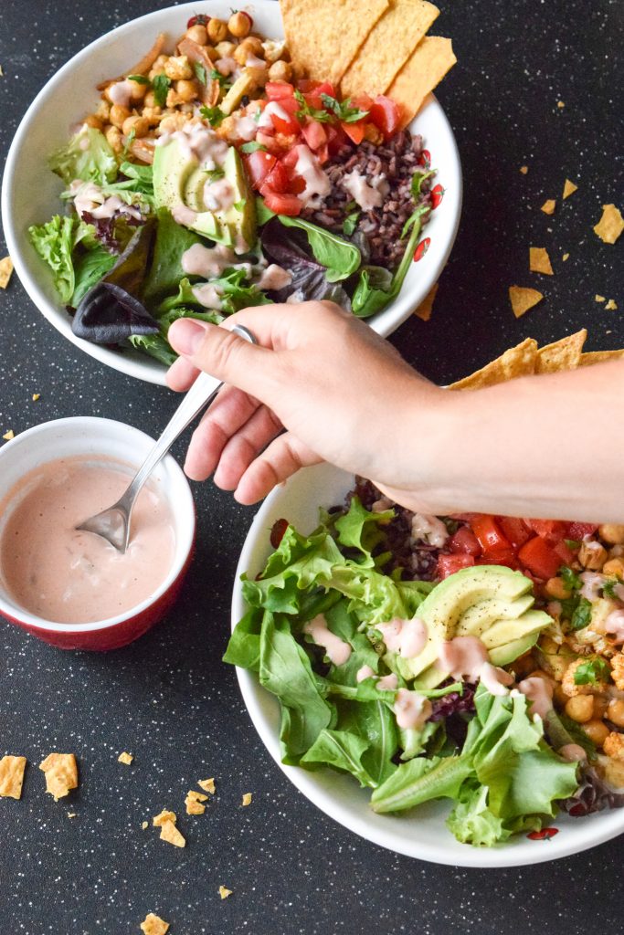 2 vegetarian taco bowls on a black backdrop, surrounded by broken tortilla chips. Arm reaches across to the left to spoon yogurt dressing.