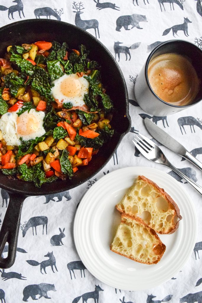 Breakfast hash, made with eggs and veggies in a cast-iron skillet, atop a white tea towel. To the side, a white plate with toast, utensils and a mug of coffee to the side.