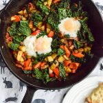 2 poached eggs and veggies in a cast-iron skillet, atop a white tea towel. To the side, a white plate with toast.