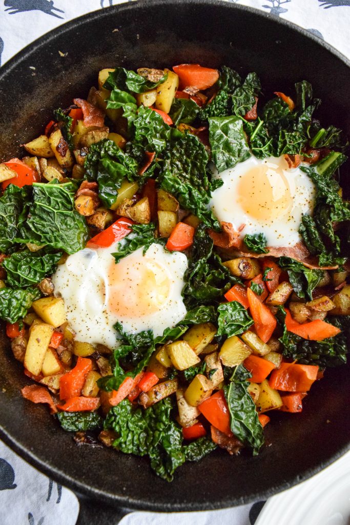 Breakfast hash of two poached eggs cooked with red peppers, kale, bacon and potatoes.