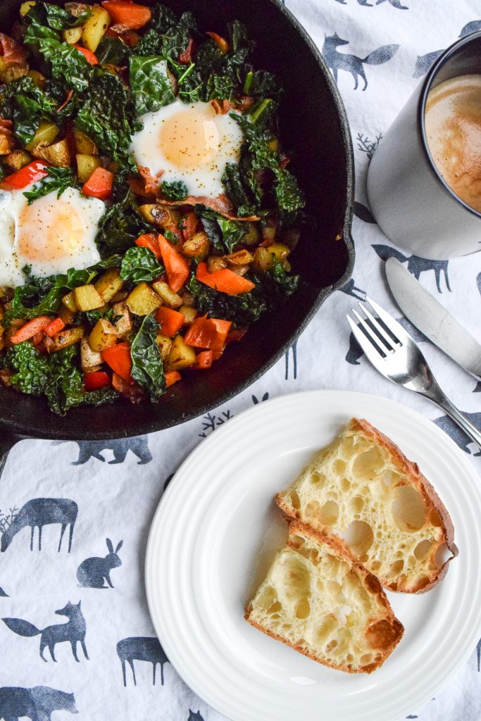 2 poached eggs and veggies in a cast-iron skillet, atop a white tea towel. To the side, a white plate with toast, utensils and a mug of coffee to the side.