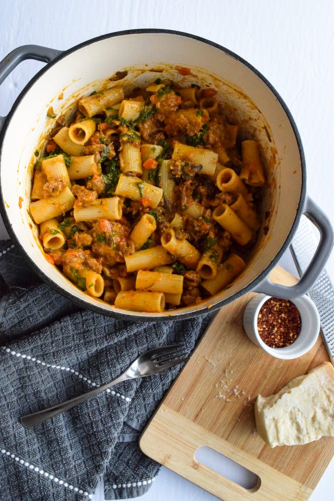 Grey dutch oven filled with rigatoni in a sausage, squash and mushroom pasta sauce. On the side, a small wooden cutting board with a piece of parmesan cheese and a small white bowl.