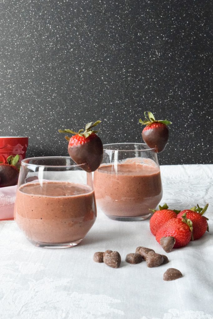 Two smoothies in short glasses, each rimmed with a chocolate covered strawberry. Extra strawberries and heart-shaped chocolate chips to the side.