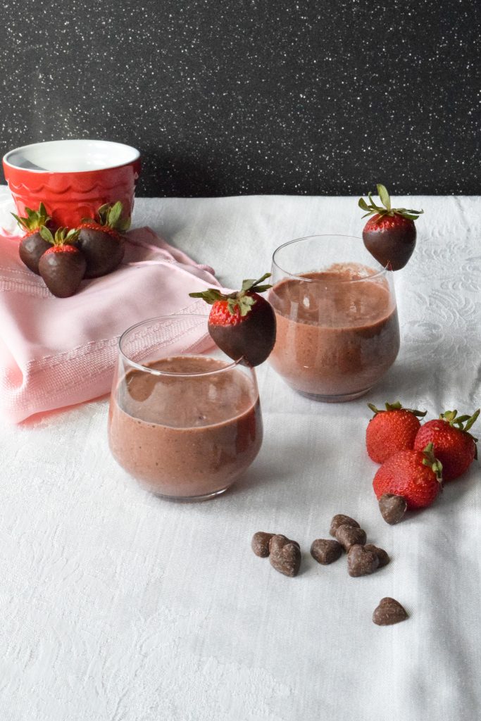 Two smoothies in short glasses, each rimmed with a chocolate covered strawberry. Extra strawberries and heart-shaped chocolate chips to the side.