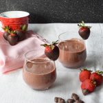 Two smoothies in short glasses, each rimmed with a chocolate covered strawberry. Extra strawberries and heart-shaped chocolate chips to the side.