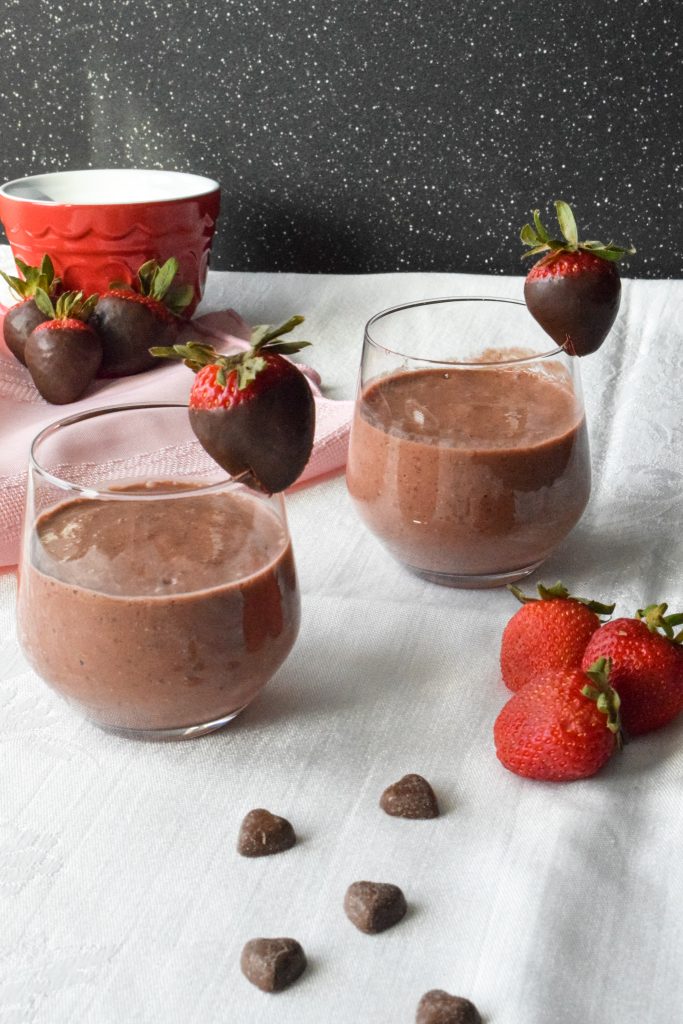 Two smoothies in short glasses, each rimmed with a chocolate covered strawberry. Extra strawberries and heart-shaped chocolate chips to the side.