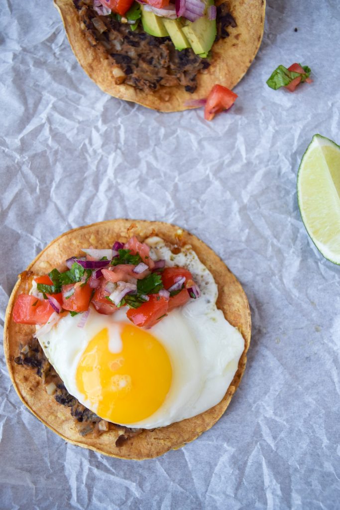 Close up shot of a black bean tostada with an egg, topped with fresh pico de gallo. On the side, another tostada and a lime wedge.